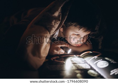 Schoolboy boy studies in the dark under the covers a book on astronomy