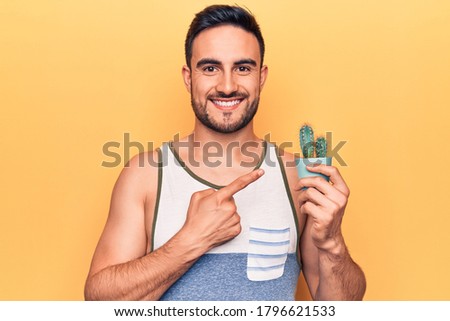 Young handsome man with beard wearing sleeveless t-shirt holding cactus plant pot smiling happy pointing with hand and finger
