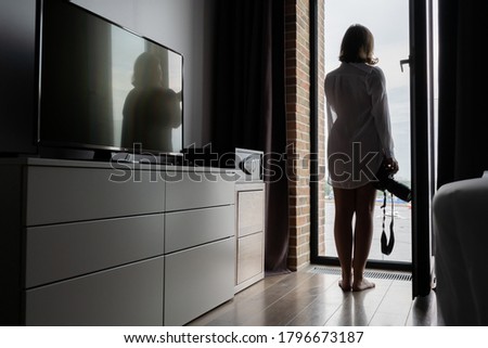 Back view of silhouette woman photographer in a transparent nightgown taking picture using camera standing near open panoramic window of apartments. 