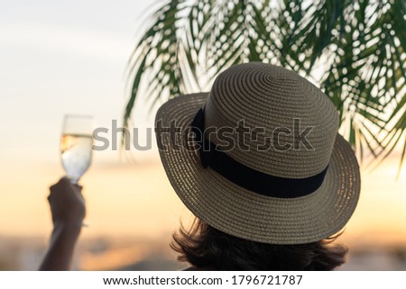 Back view of lonely beautiful girl with glass of champagn  in a straw hat against the background of the sea in branches of palm trees. Sunset beach. Summer and freedom concept.