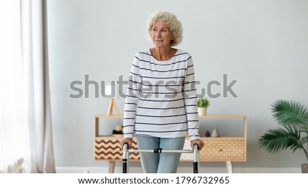 Happy middle aged senior hoary retired woman using walking frame, enjoying rehabilitation procedures indoors. Smiling older disabled grandmother making steps with walker, lifestyle. Royalty-Free Stock Photo #1796732965