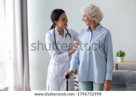 Smiling young physiotherapist cuddling shoulders of happy old retired woman leaning on wooden walking cane. Pleasant female nurse in white coat starting rehabilitation procedure with elderly patient. Royalty-Free Stock Photo #1796732998