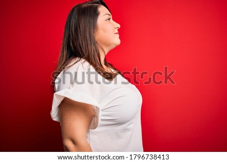Beautiful brunette plus size woman wearing casual t-shirt over isolated red background looking to side, relax profile pose with natural face with confident smile.