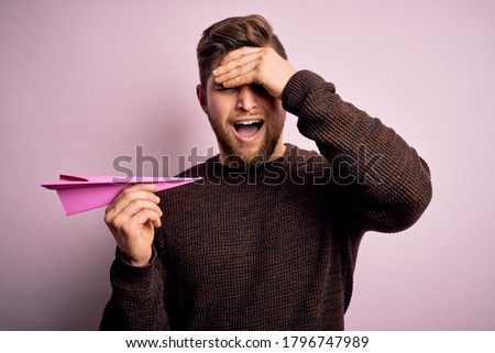 Young blond man with beard and blue eyes holding pink paper plane over isolated background stressed with hand on head, shocked with shame and surprise face, angry and frustrated. Fear and upset