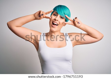 Young beautiful woman with blue fashion hair wearing casual t-shirt over white background Doing peace symbol with fingers over face, smiling cheerful showing victory