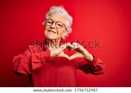 Senior beautiful grey-haired woman wearing casual shirt and glasses over red background smiling in love doing heart symbol shape with hands. Romantic concept.