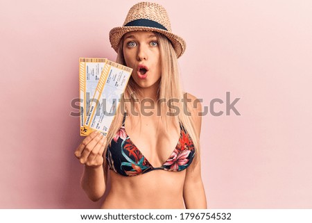 Young beautiful blonde woman wearing bikini and hat holding boarding pass scared and amazed with open mouth for surprise, disbelief face 