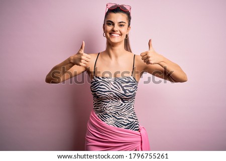 Young beautiful brunette woman on vacation wearing swimsuit over pink background success sign doing positive gesture with hand, thumbs up smiling and happy. Cheerful expression and winner gesture.