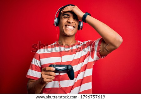 Handsome african american gamer man playing video game using jostick and headphones stressed with hand on head, shocked with shame and surprise face, angry and frustrated. Fear and upset for mistake.