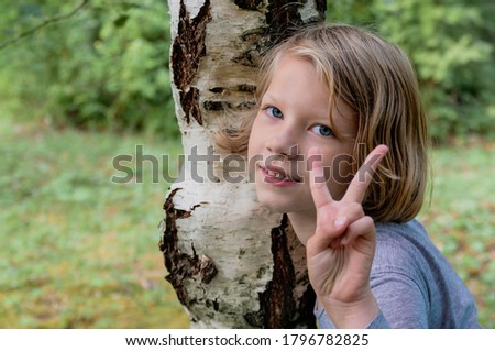 Portrait of a cute seven-year-old girl showing the victory sign standing next to a birch tree