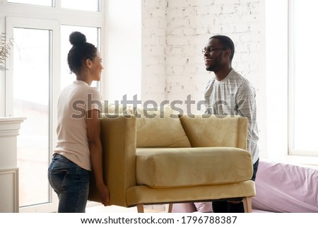 Moving day. Happy loving millennial african spouses furnishing new house or apartment, smiling black husband and wife carry new armchair to living room, young married couple making home improvements Royalty-Free Stock Photo #1796788387