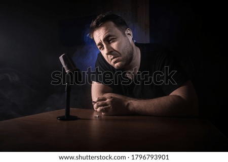 Caucasian man news broadcaster sat at a desk with microphone in dark room. Performer with microphone.