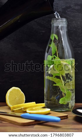 Hot water is poured into a glass bottle containing slices of lemon and mint - homemade tea as refreshment in summer with mint and lemon on one surface - background with homemade tea