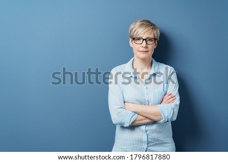 Portrait of mature blonde woman wearing glasses standing against blue background with arms crossed Royalty-Free Stock Photo #1796817880