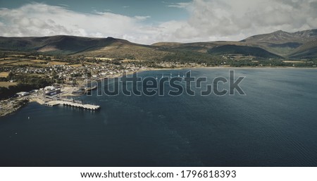 Scotland ocean port aerial view: ship, yachts and boats on coastal water of Atlantic gulf. Scottish cityscape with homes, cottages, roads against greenery hills. Cinematic shot