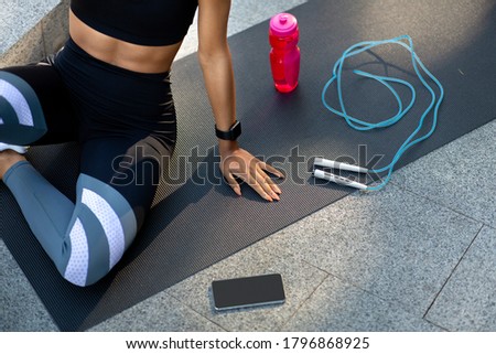 Sporty girl resting on fitness map, having skipping rope, bottle with water and smartphone with blank screen, training outdoor, top view, cropped Royalty-Free Stock Photo #1796868925