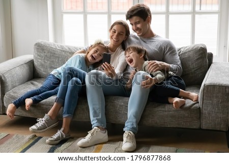 Smiling loving young couple parents showing funny video or watching childish cartoons on smartphone with laughing adorable kids siblings, enjoying stress free leisure time together on sofa at home.