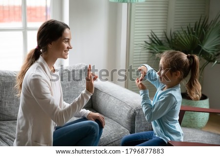 Smiling young mixed race woman teaching little cute girl sign language, showing symbols with fingers. Smiling hearing loss deaf disabled child communicating nonverbal with mommy in living room. Royalty-Free Stock Photo #1796877883