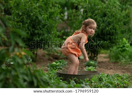 
A little girl with blond hair stands in a bath with water and splashes on the green grass. The kid is having fun, splashing and laughing. Happy child swimming outdoors in the spring garden. Summer, h