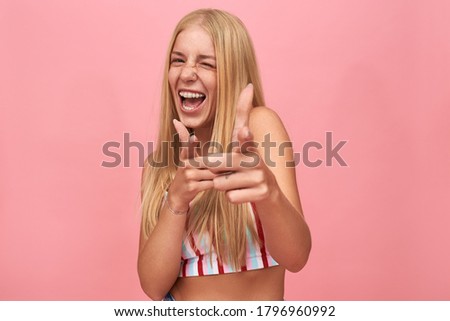 Cheerful excited hipster girl with loose straight hair winking, pointing fore fingers at camera, being in good mood. Stylish young Caucasian woman having fun, making gesture. Body language