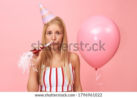 Isolated image of upset sad young European female wearing conical hat and summer crop top celebrating birthday, having upset facial expressing blowing party horn, holding pink helium balloon