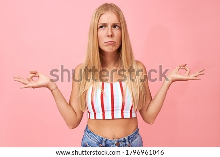 Beautiful young Caucasian female making grimace while doing meditation, having skeptical facial expression, trying to relax mind and body, looking for balance within. Peace, zen and harmony