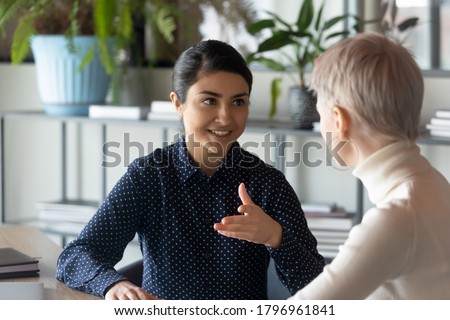 Friendly millennial indian businesswoman talking to blonde female colleague, sitting at table in office. Two young multiracial employees discussing working issues or enjoying informal conversation. Royalty-Free Stock Photo #1796961841