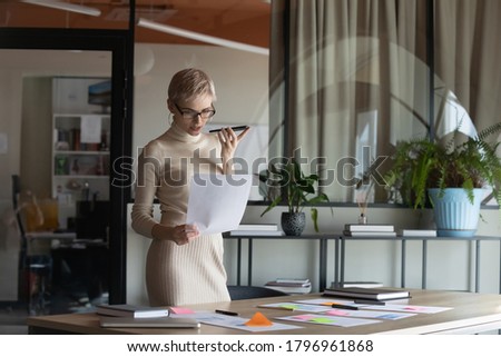 Concentrated young female blonde ceo manager using virtual assistant on phone, checking information in financial paper document, 30s businesswoman leader sending voice mail for client in modern office