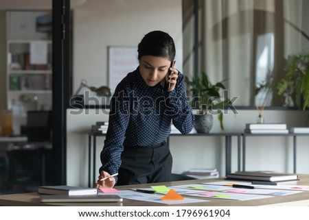 Concentrated serious young indian female employee managing working tasks with colorful stickers or preparing presentation, holding mobile phone call with boss or consulting client distantly in office. Royalty-Free Stock Photo #1796961904