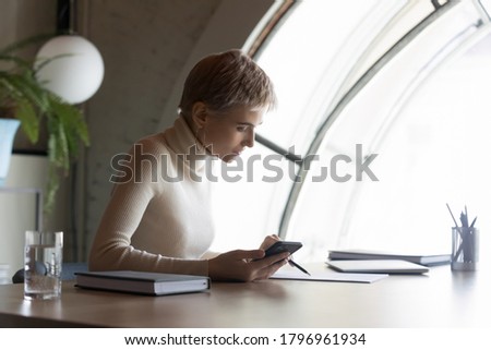 Side view confident serious 30s short haired female notary reading paper business agreement, checking newest law in mobile phone application, concentrated editor working on handwritten documents.