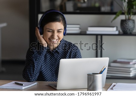 Happy positive young indian businesswoman wearing wireless headphones, listening favorite energetic audio music, relaxing in office, smiling employee enjoying funny educational online lecture. Royalty-Free Stock Photo #1796961997