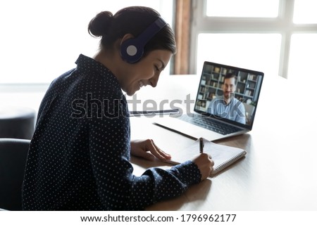 Smiling young indian woman in headphones learning practicing foreign language with confident male tutor distantly on computer. Happy mixed race girl listening to educational webinar, writing notes. Royalty-Free Stock Photo #1796962177