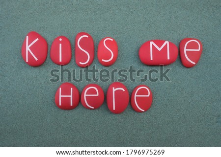 Kiss me here, creative love message composed with red colored stone letters over green sand