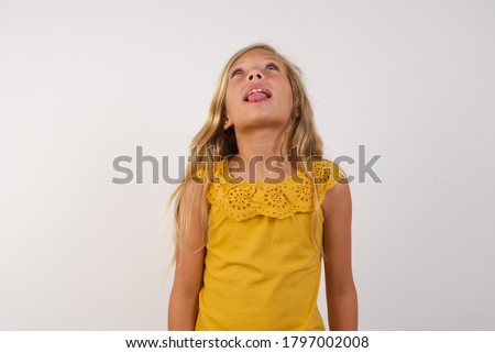 Little girl with beautiful blonde hair over white background showing grimace face crossing her eyes and showing tongue . Being funny and crazy