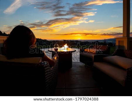 A woman relaxes with a glass of wine at night in front of an outdoor firepit on a patio of a luxury home overlooking a city and valley