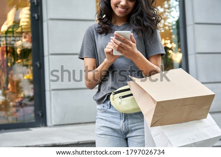 Smiling young African American ethnic girl shopper customer standing on city street holding shopping bags and smart cell phone using mobile fashion sale ecommerce app near shop mall store, closeup.