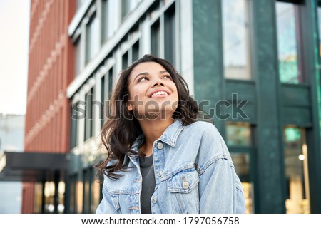 Confident happy beautiful young hipster African American woman wearing denim jacket looking up standing on city street outdoors dreaming, thinking or good future on urban buildings background. Royalty-Free Stock Photo #1797056758