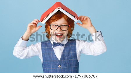 Delighted boy in glasses smiling for camera and putting book on head while having fun during school studies against blue background

