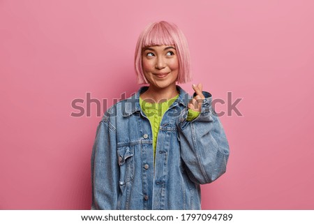 Horizontal shot of dreamy thoughtful young Asian woman makes love sign, shapes mini heart with fingers, feels happy, wears stylish denim clothes, pink background. Body language, korean style Royalty-Free Stock Photo #1797094789