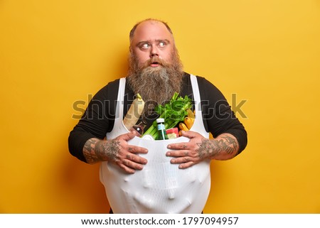 Obese greedy man stocks foodstuff, eats too much, suffers from overweight and gluttony, keeps hands on belly full of products, has tattooed arms, thick beard, concentrated with frightened look