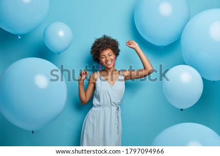 Positive smiling dark skinned woman dances carefree, keeps arms raised, wears blue fashionable dress, closes eyes, spends free time on disco party, moves against blue background with balloons