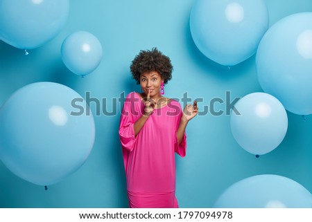 Happy mysterious dark skinned Afro American woman makes silence gesture, asks to be quiet, dressed in pink long dress, spreads rumors, poses against blue background with inflated balloons around.