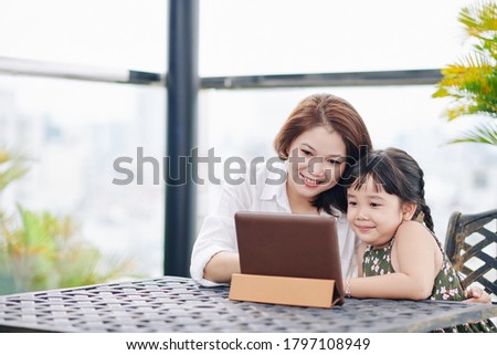 Smiling Asian mother and daughter sitting at table outdoors and watching cartoon on digital tablet