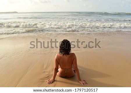A young caucasian girl is sitting in the beach with her back to the camera and observes the wide ocean and the crashing waves on a sunny summer day.