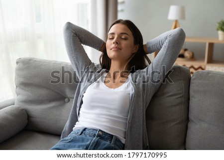 Young beautiful peaceful brunette woman leaning on comfortable sofa, stretching back with folded hands behind head, daydreaming with closed eyes, enjoying calm carefree moment alone in living room. Royalty-Free Stock Photo #1797170995