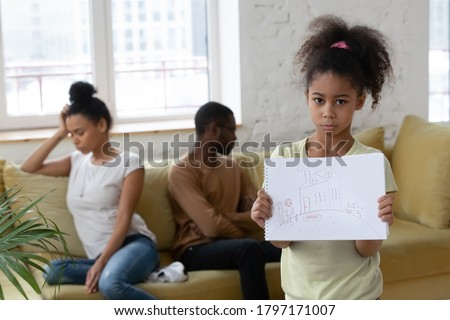 Sad unhappy little african kid girl suffering, feeling bad and lonely looking at camera holding drawing of happy family while her parents having conflict or divorcing.