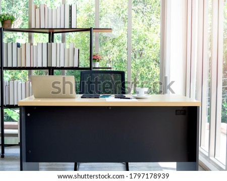 Modern office interior. Well-lit contemporary workplace with equipment, laptop computer and coffee cup on desk and chair with bookshelf in the back near large window glass overlooking the tree. Royalty-Free Stock Photo #1797178939