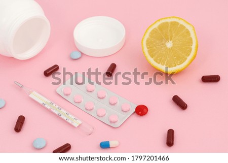 Multi-colored tablets in capsules on a light pink background. Thermometer, lemon and a white container for tablets. 