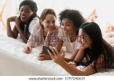 Multi-ethnic best friends lying in bed. Asian ethnicity girl holding smartphone showing to intimates new app, photos social network account. Women having fun watching videos on-line. Pajama party