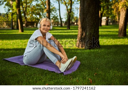Charming lady sitting on yoga mat and smiling while enjoying favorite songs outdoors